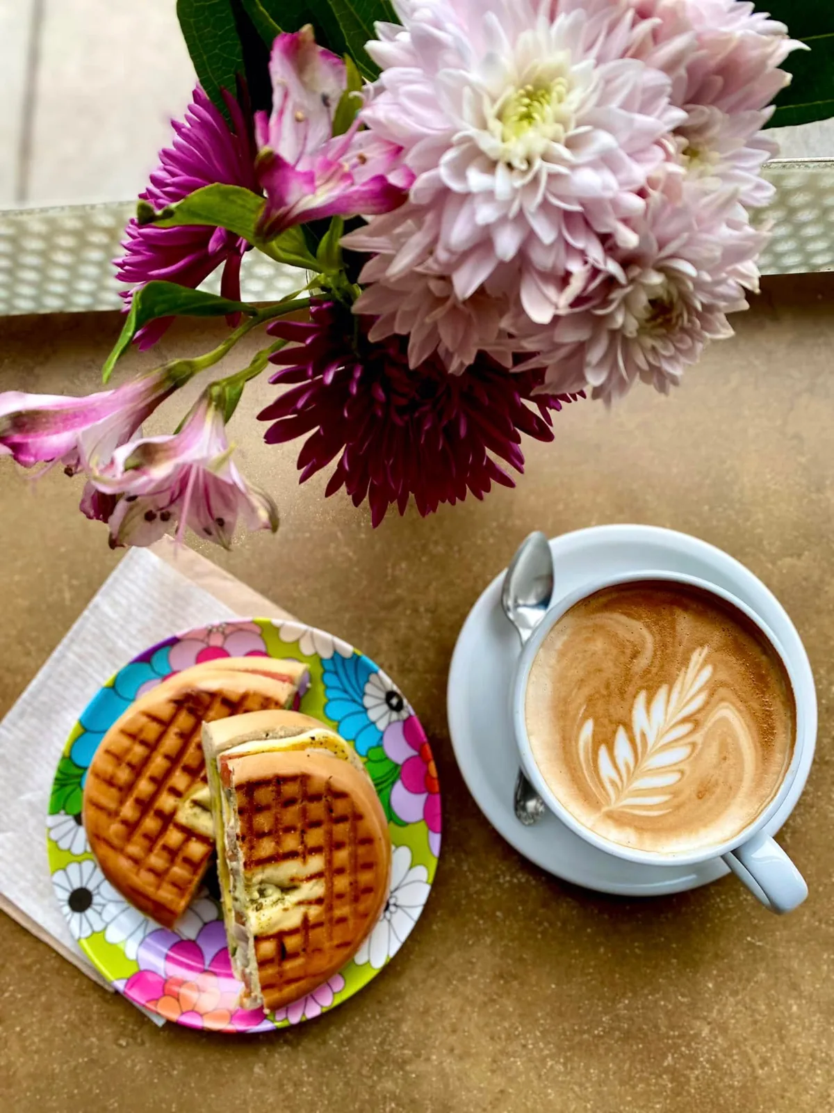 A cappuccino with leaf latte art beside waffle sandwiches and a vase of flowers