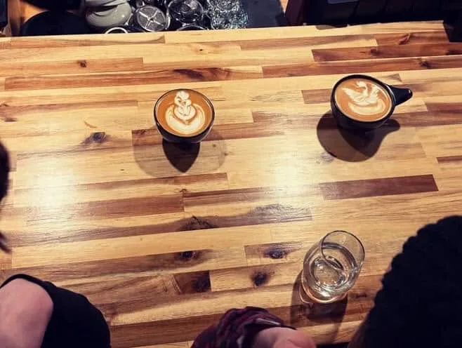 Two cappuccinos with latte art and a glass of water on a butcher-block table