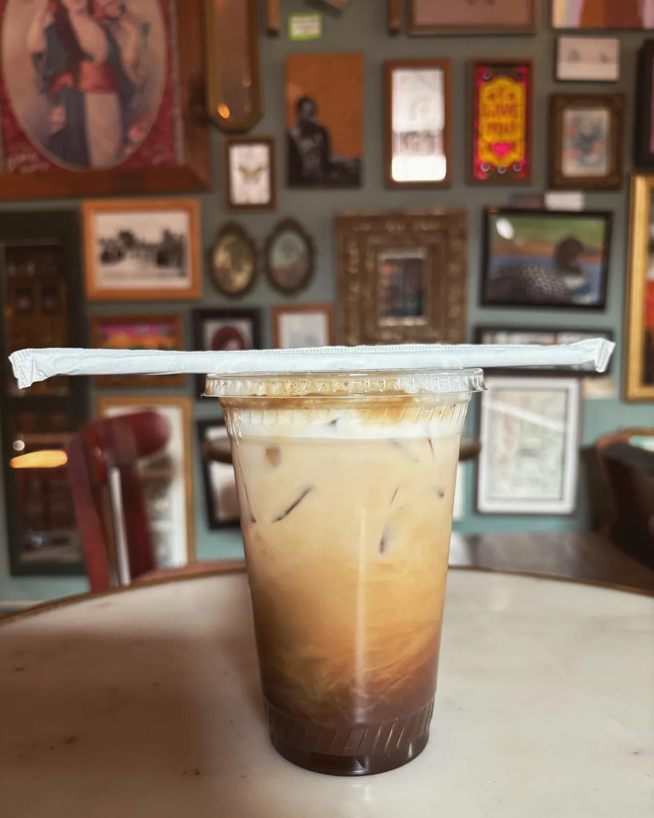 An iced latte on a table in front of a wall hung with framed pictures