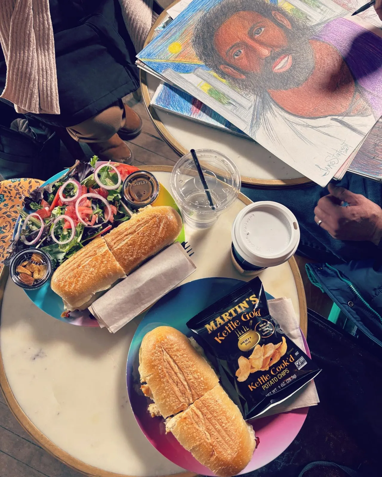A sub sandwich, a salad and iced drinks on a table, with someone sketching beside them